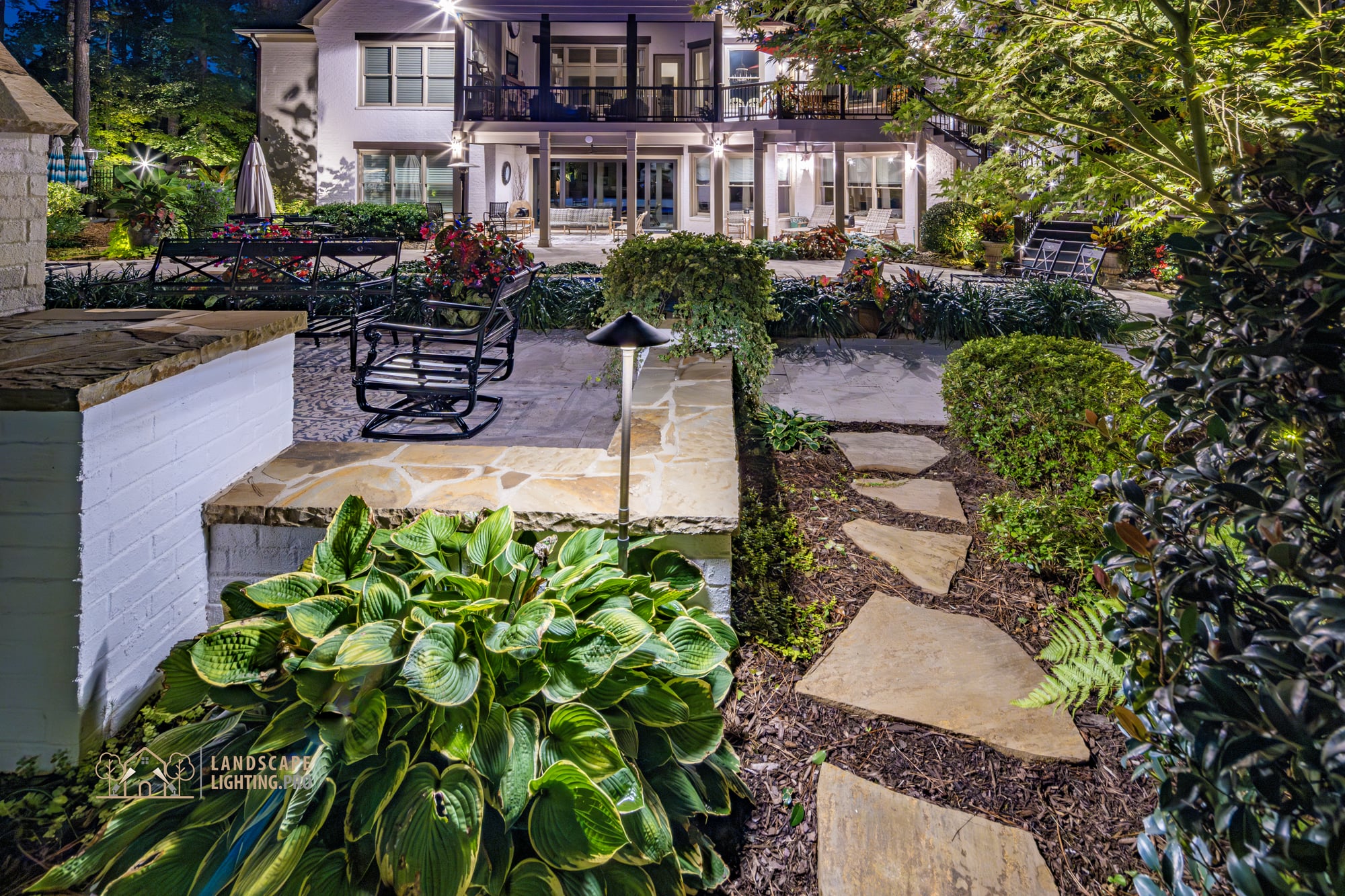 The rear of the home from the garden side, with a flagstone approach, balcony, and manicured shrubs at dusk