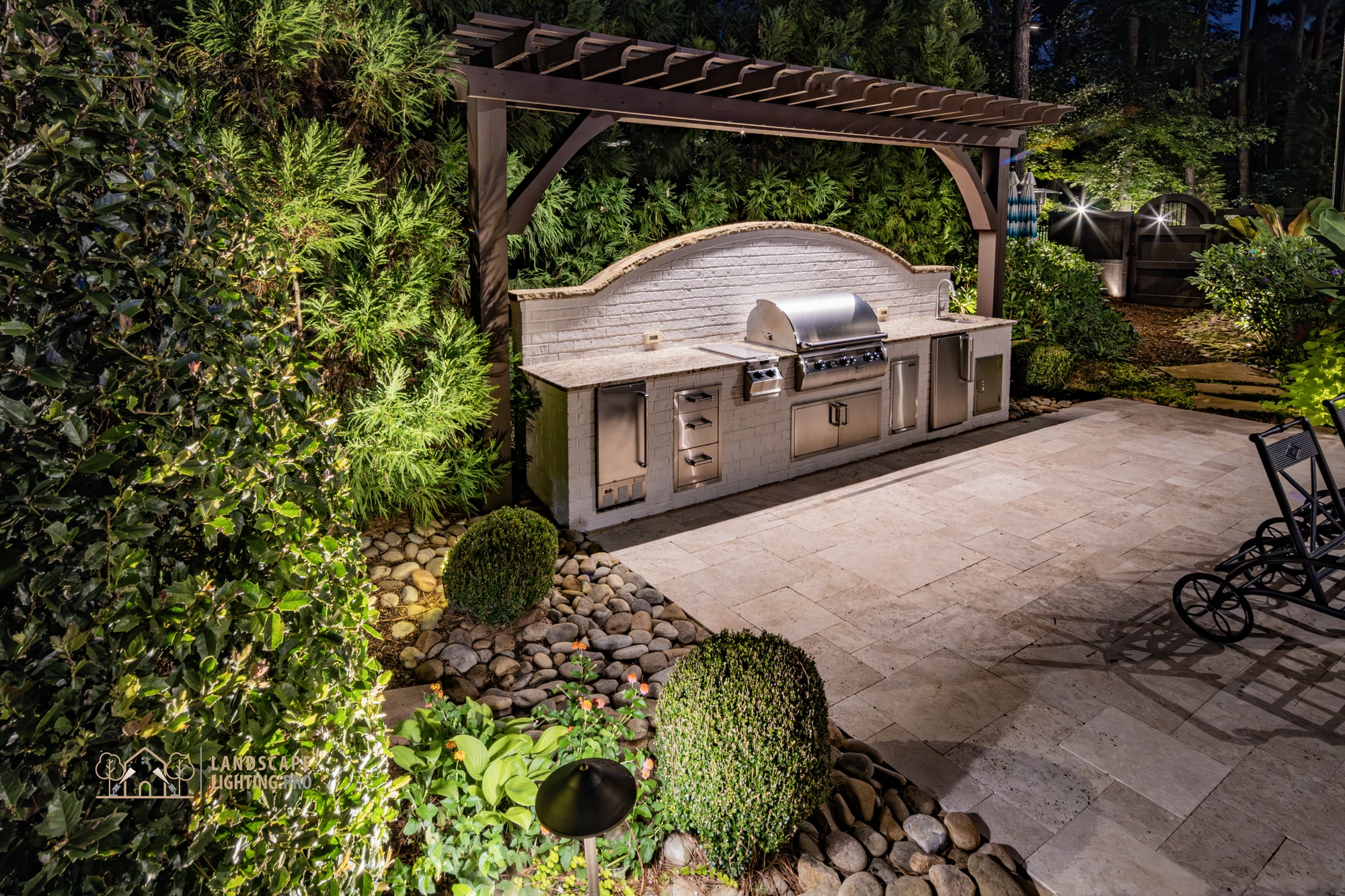 An outdoor grill station under a timber pergola, framed by evergreen plantings and travertine paving at night