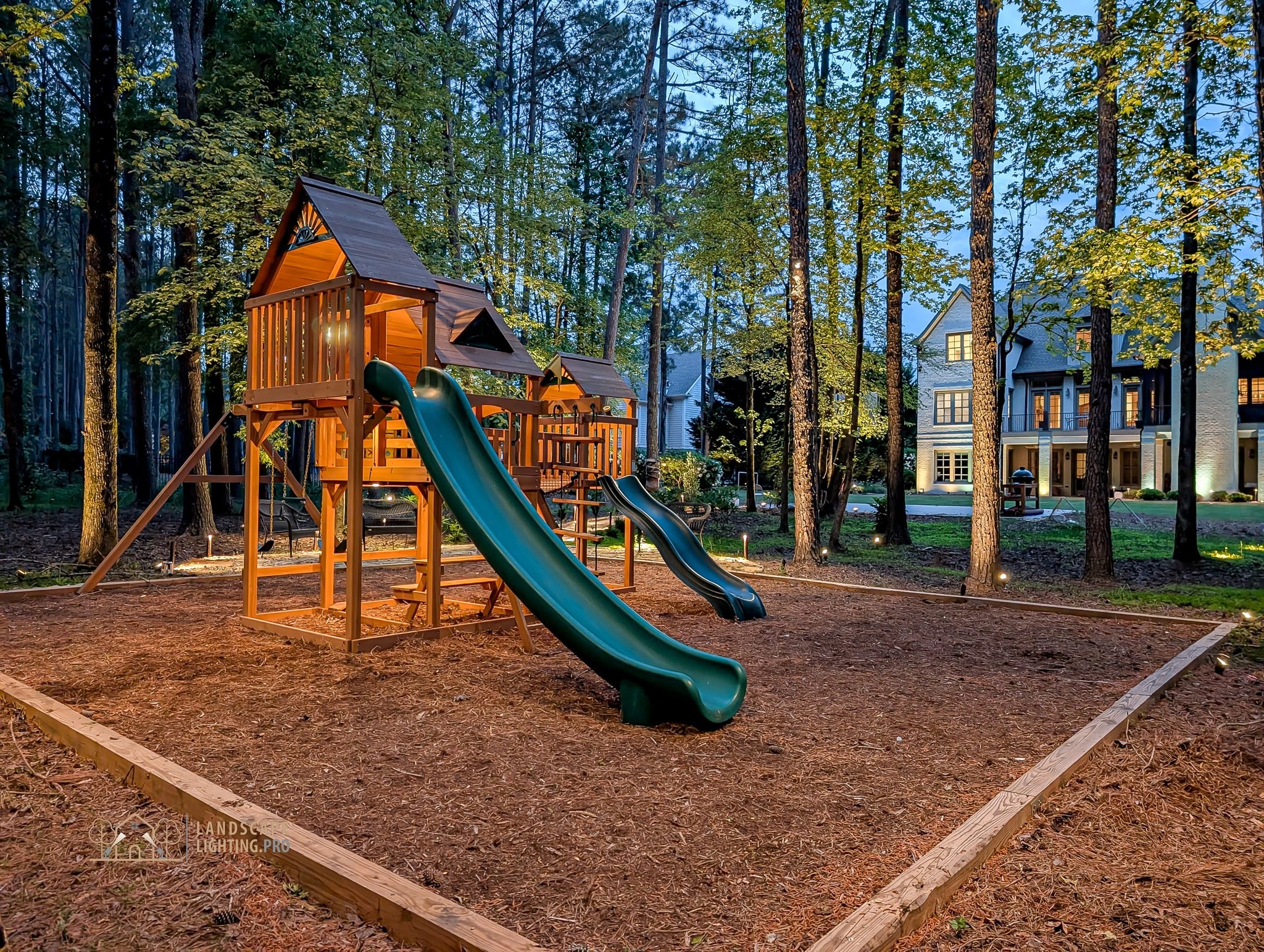 A wooden playset with a green slide set into a pine grove and lit at dusk