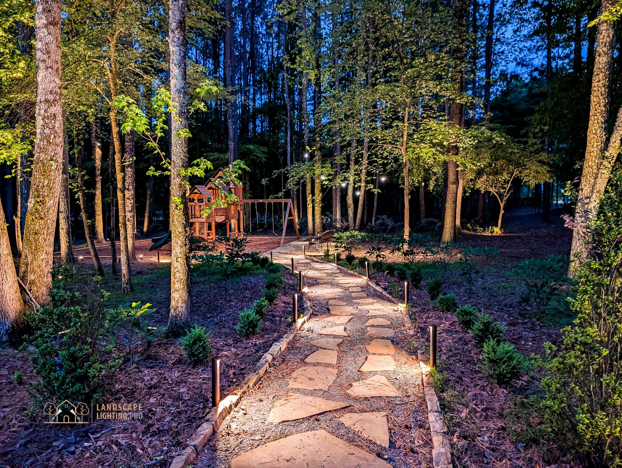 A flagstone path lit at grade running through a pine grove toward a wooden playset at dusk