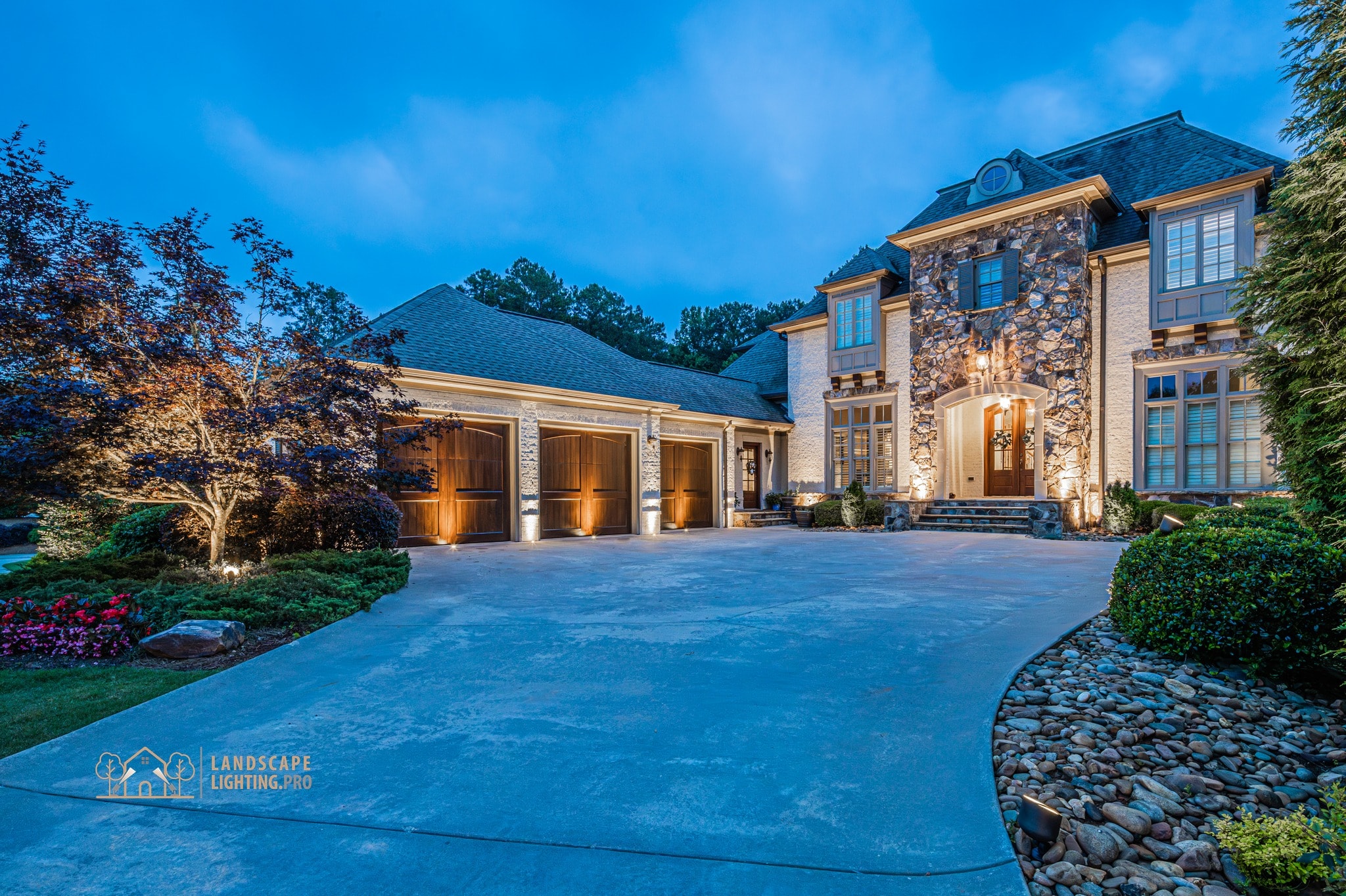 A stone-and-brick estate home at dusk with illuminated façade, copper-trimmed garage doors, and a paved circular driveway